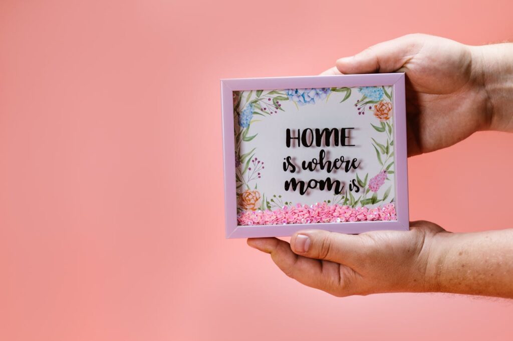Hands holding a floral shadow box with 'Home is where mom is' text, perfect for Mother's Day gifting.